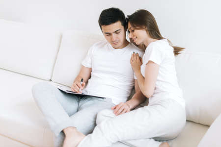 Pretty guy and beautiful girl in light clothes sit on white sofa in bright modern living room and discuss joint plans for future. Young man writes notes with pen in notebook. Girl hugs his boyfriendの写真素材