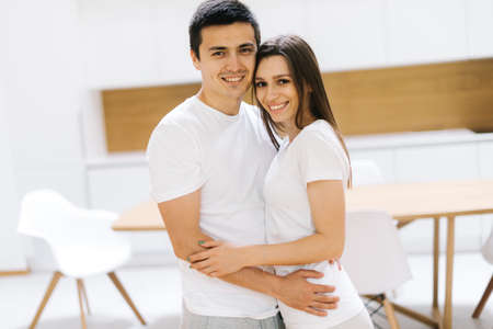 Beautiful young couple is hugging, looking at camera and smiling on white bright background. Handsome girl and cute guy with white T-shirtsの写真素材