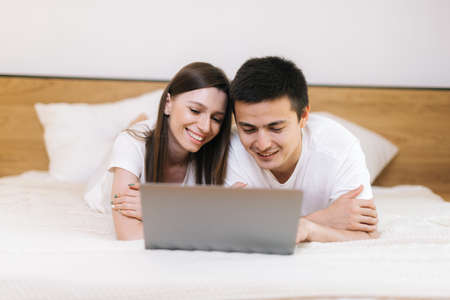 Young couple relaxing in light bed with laptop. Lying together in light bedroom. Happy smiling young couple in love at home, laying in bed, looking at laptop computerの写真素材