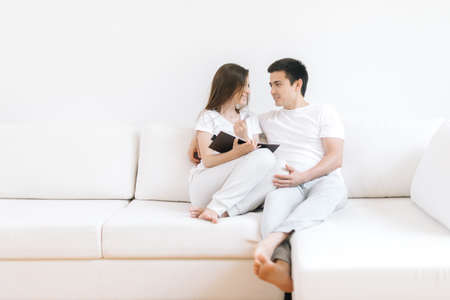 Loving couple hugging holding hands relaxing on cozy sofa. Smiling boyfriend and girlfriend looking at each other embracing at couch. Young happy lovers resting enjoying weekend at home togetherの写真素材