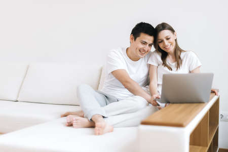 Young married couple sitting on soft sofa and looking at laptop in bright living room. Happy people buying things for house in online store sitting in light apartment. Guy and girl smiling.の写真素材