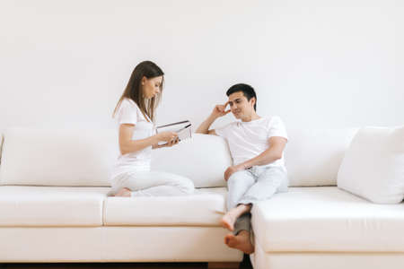 Couple is reading book at house in white living room. Happy couple reading mail and checking accountancy looking each other sitting on a sofa at homeの写真素材