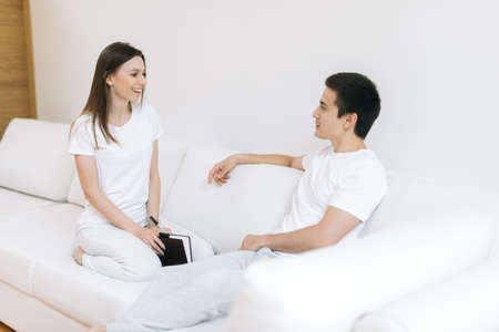 Young happy couple sitting on white sofa, smiling at each other. Girl holds black notebook in her hands. Guy looks at his girlfriend with his eyes in loveの写真素材