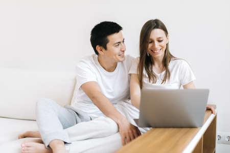 Portrait of happy young couple sitting on couch and working on laptop at home. Happy young couple is working on laptop in bright apartment.の写真素材