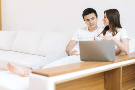 Young woman showing important information to her boyfriend on the laptop. Couple at home. Beautiful young couple is using a laptop, hugging and smiling while sitting on couch at homeの写真素材