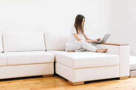 Young business woman looking at laptop computer at home. Female person using computer for online work on white couch. Happy girl looking at notebook screen sitting on sofa. Home work conceptの写真素材