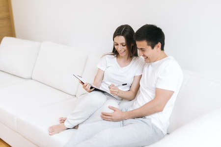 Pretty guy and beautiful girl in light clothes sit on white soft couch in modern living room and discuss joint plans for future. Young woman writes notes with pen in notebook. Guy hugs his girlfriendの写真素材
