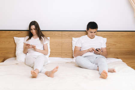 Young couple of guy and girl actively using phones. Two people totally focused on their smartphones sitting on bed at homeの写真素材