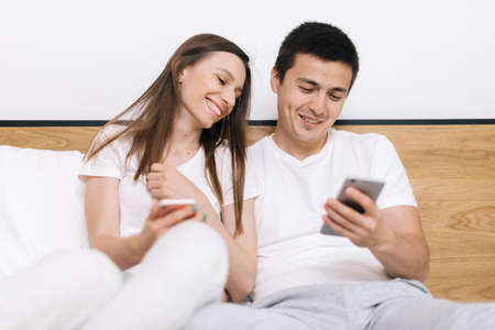 Attractive young married couple is sitting on sofa in living room and laughs watching video on smartphone. Portrait of happy loving beautiful couple sitting on bed in bright modern apartmentの写真素材
