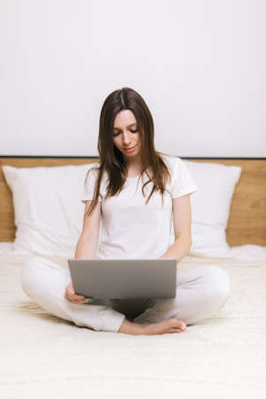 Young woman typing on laptop sitting on sofa. Girl freelancer working on laptop in home. Female using notebook in modern white living room. Student performs task. Female thought about solving problemの写真素材