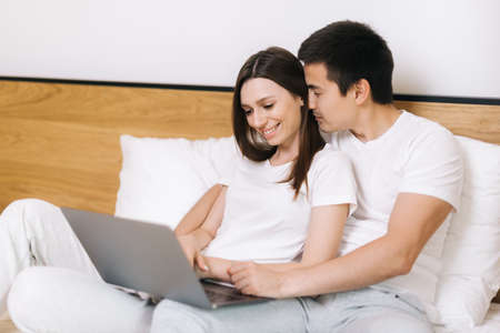 Happy young couple of guy and girl is lying in bed and watching film on laptop. Young married couple lying on bed at home and using laptop. Happy couple is lounging in bed before going to bedの写真素材