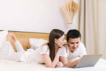 Young happy caucasian young man and woman lying on the bed, talking and using laptop in bright bedroomの写真素材