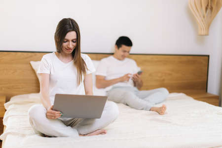 Girl working on laptop sitting in bed. Guy uses phone behind his girlfriend. Man and a woman using mobile phone and laptop while sitting in bed. Girl is typing on notepad, the guy is surfing Internetの写真素材