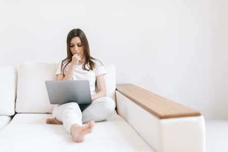 Young woman with laptop on light couch. Girl freelancer working on laptop in home. Female using notebook in her modern white living room. Student performs task. Female thought about solving problemの写真素材