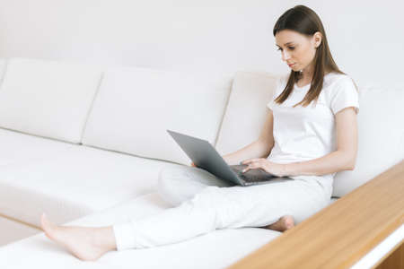 Serious young woman think on ideas for development of own website sitting on soft comfortable sofa in light home. Girl blogger typing on laptop computer in bright modern living roomの写真素材