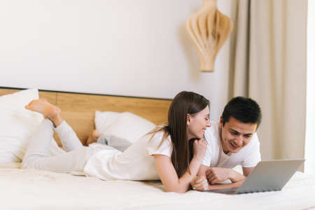 Close-up portrait of young handsome couple lying on bed in the evening and watching something funny on the laptop computer in bright bedroomの写真素材