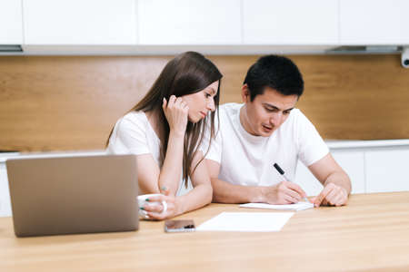 Young couple is talking on kitchen and write in notepad entry note. Confident man explaining to girl information and writes it on paper. Woman smiling listening to partner. Laptop standing on tableの写真素材