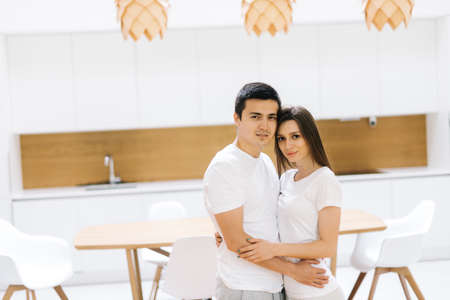 Happy smiling couple is hugging each other and looking at camera. Nice stylish couple hugging each other and looking at camera on light white background. Kitchen chairs and table in backdropの写真素材