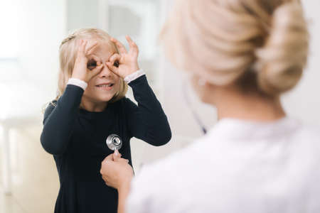 Cute funny kid girl with blond hair fooling around when pediatrician with stethoscope is examining her. Cute little kid girl in hospital at doctor appointment.の写真素材