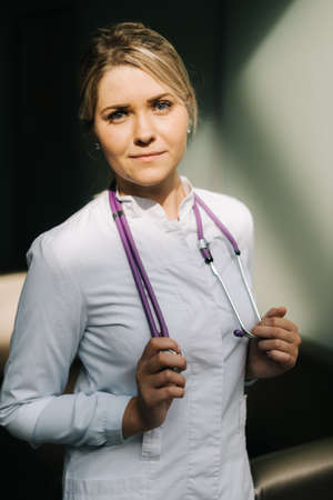 Portrait of young handsome woman doctor in white uniform standing with stethoscope. Girl looking at cameraの写真素材
