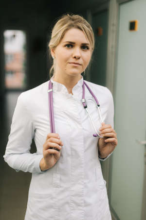 Portrait of young woman doctor wearing white uniform standing in hospital corridor. Female medical nurse or practitioner with stethoscope looking at camera. Healthcare conceptの写真素材