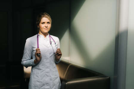 Portrait of young handsome woman doctor in white uniform standing with stethoscope. Girl looking at...