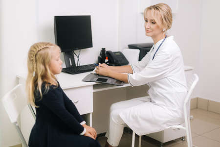 Cute blonde kid girl patient sit on chair talking to woman doctor. Young woman doctor pediatrician talking to kid girl patient at medical checkup appointmentの写真素材