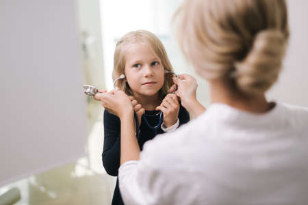 Cute funny kid girl with blond hair wears a stethoscope under supervision of pediatrician. Cute little kid girl in hospital at doctor appointment.の写真素材