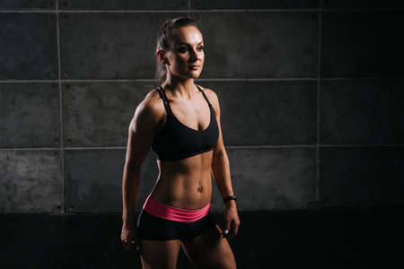 Muscular young woman with perfect athletic body in sports black bra is standing and posing on dark isolated backgroundの写真素材