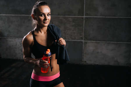 Young muscular woman bodybuilder with perfect athletic body in black sportswear with towel on shoulder, is holding bottled water in hand on isolated dark background. Sporty girl looking at the cameraの写真素材