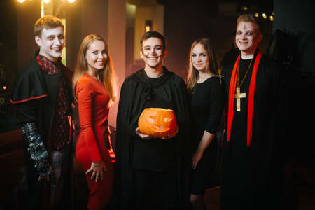 Young people in costumes are celebrating Halloween. Group of young cheerful guys and girls dressed in Halloween clothes are looking at camera and posing with a pumpkin in room with black and red wallsの写真素材