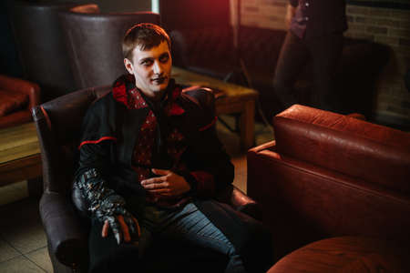 Young man in a Halloween costume of Count Dracula with colorful make-up on his face sits behind a chair and looks at the cameraの写真素材