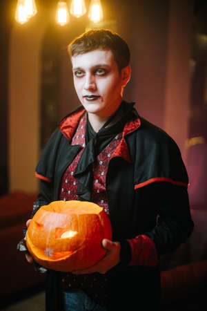 Guy in a Halloween costume of Count Dracula holds a carved pumpkin in his hand and looks into the camera.の写真素材
