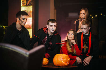 Group of five friends are celebrating Halloween in a cafe. Guys dressed as scary holiday monsters and dressed in colorful costumes. Holiday carved pumpkin on tableの写真素材