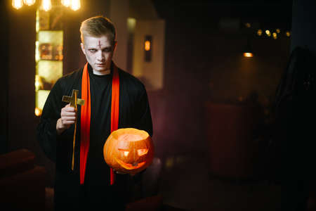 Guy as a priest holds a cross and a Halloween pumpkin. Young man looks into the camera.の写真素材