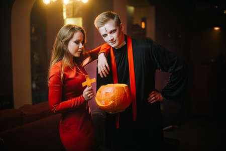 Young man in a Halloween priests costume holds a carved pumpkin in his hand and looks at the camera. A girl in a red dress leans on the guys shoulderの写真素材