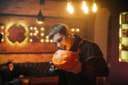 Young man in a Halloween costume of Count Dracula holds a carved festive pumpkin in his hand and ominously tries to bite her.の写真素材