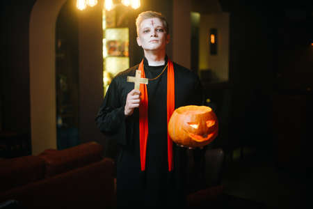 Young man in a Halloween priest costume holds a carved pumpkin and a religious cross. Guy looks into the cameraの写真素材