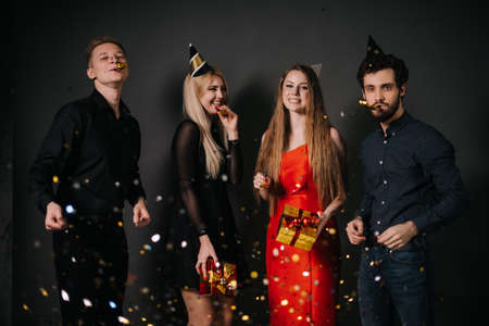 Two cheerful guys blow a festive whistle and throw up a colorful confetti. Two beautiful happy girl are holding bright gift boxes in hands. Shooting in on isolated dark background.の写真素材