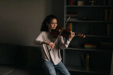 Beautiful young woman violinist plays the violin at home, sitting on the couchの写真素材