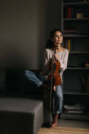 Beautiful young woman posing with a violin at home sitting on the couch. Girl smiles and looks awayの写真素材