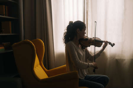 Beautiful girl musician plays on the background of a window at home, sitting on a soft chairの写真素材