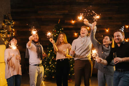 Young cheerful friends dance with Bengali lights, have fun and carefree time celebrating New Year. People dancing with sparklers on dark room and clinking with champagne glasses.の写真素材