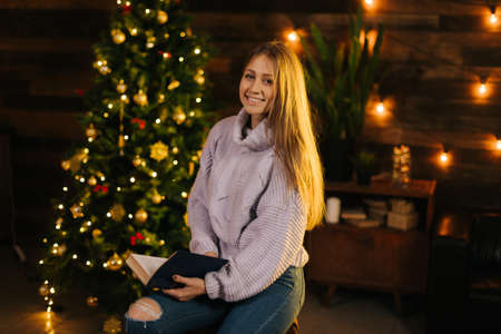 Beautiful girl in cozy sweater posing with a book in her hand against the background of a Christmas tree and wall with a festive garland. Nice girl looking at the camera.の写真素材