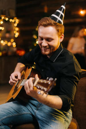 Portrait of young cheerful man in the festive hat playing on the guitar against the background of talking friends. Christmas tree with garland and wall with festive illumination in background.の写真素材