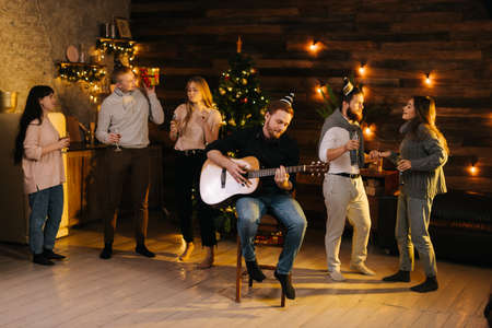Group of young man and woman sings at a Christmas party in a cozy house. Happy young man plays guitar. Christmas tree with garland and wall with festive illumination in background.の写真素材