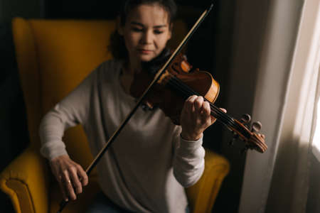 Beautiful girl musician playing at home, sitting on a soft chair. Young violinist woman sitting by the window.の写真素材