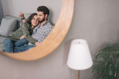 Portrait of young cute couple sitting on the couch in living room. Reflection of the hugging couple...