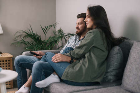 Lovely happy couple is watching TV together sitting on soft gray couch. Daily life of a male and...