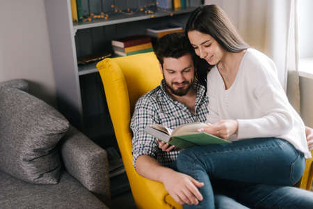 Lovely positive young couple reading a book together, sitting on a soft yellow chair. Daily life of a male and female in apartment.の写真素材